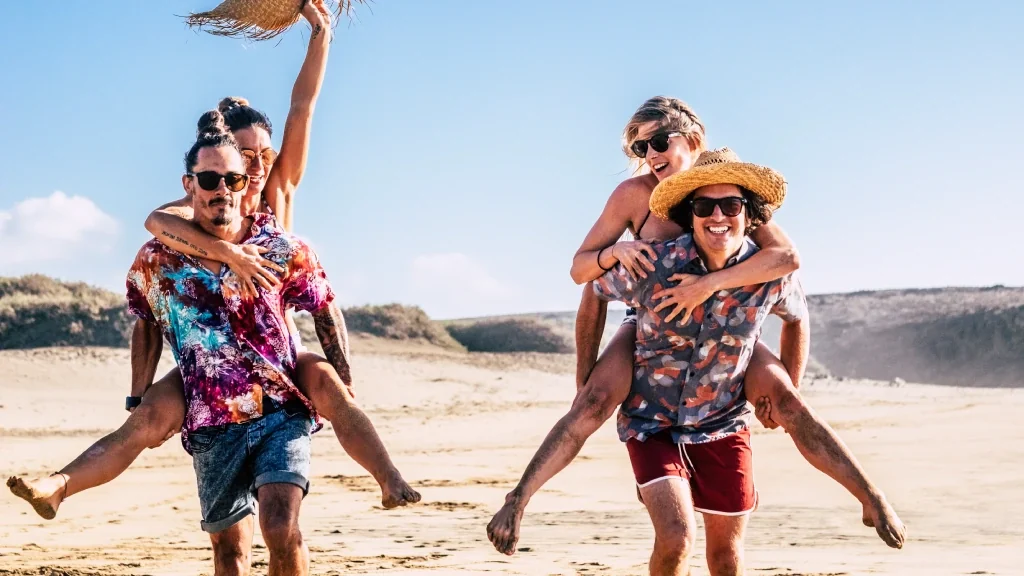 Quatro amigos rindo e brincando na areia da praia em dia de sol, dois deles carregando os outros nas costas