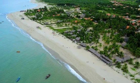 Vista aérea de praia de areia clara com coqueiros, pousadas e barcos no mar verde-azulado