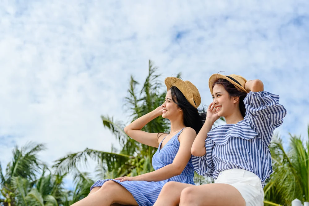 Duas viajantes de chapéu de palha sentadas ao ar livre, sorrindo, com coqueiros e céu nublado ao fundo