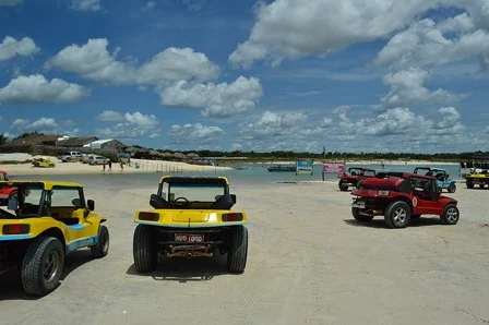 Buggies amarelos e vermelhos estacionados na areia à beira de uma lagoa, com barracas ao fundo