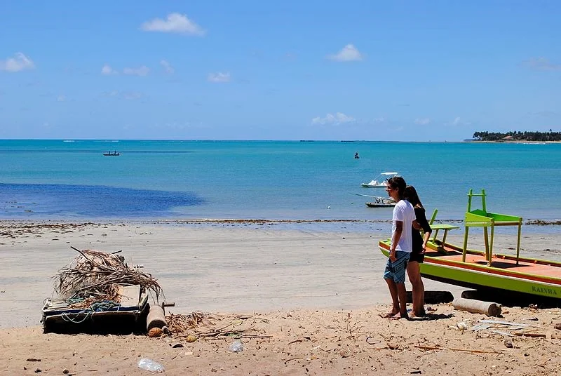 Casal na areia ao lado de um barco verde, com mar azul-turquesa e lanchas ao fundo