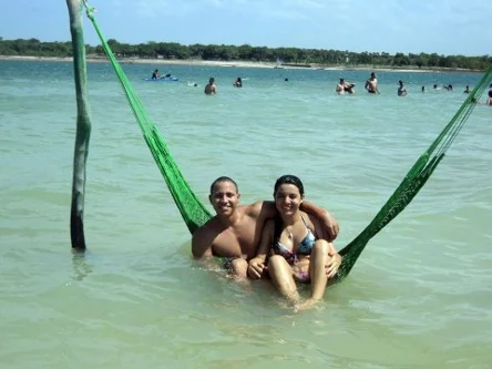 Casal sentado em rede verde armada dentro da água rasa de uma lagoa, com banhistas ao fundo
