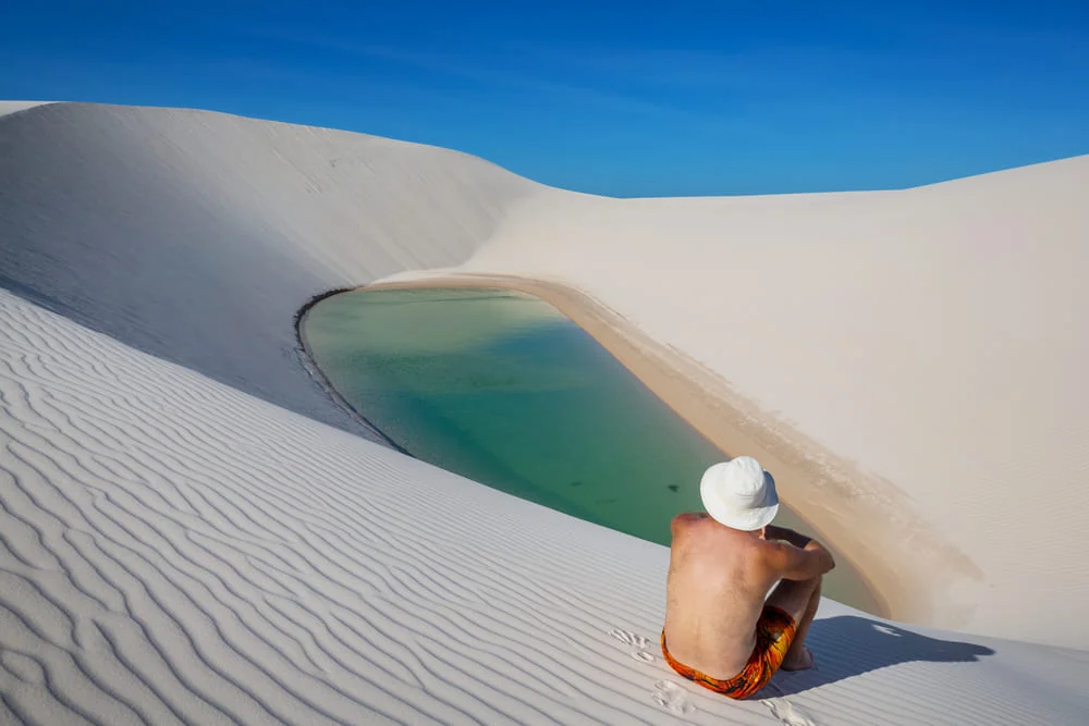Homem de chapéu branco sentado em duna de areia branca diante de uma lagoa verde-esmeralda, sob céu azul