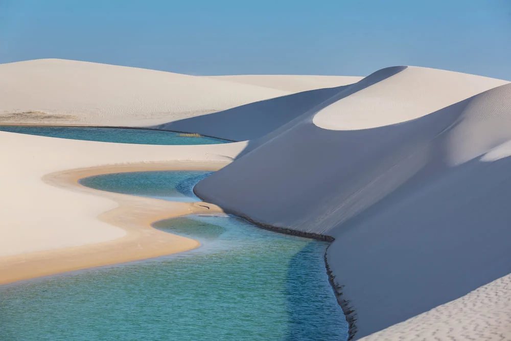 Dunas de areia branca contornando lagoas de água azul-esverdeada, com faixa de areia dourada na margem e céu azul