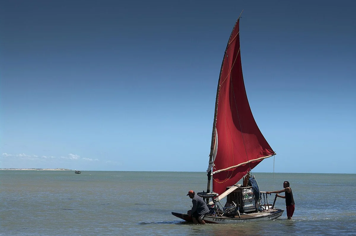 Jangada de vela vermelha com dois pescadores a bordo, navegando em mar raso perto da faixa de areia