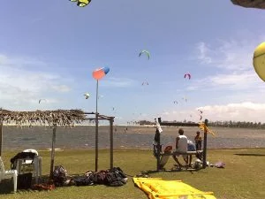 Pipas coloridas de kitesurf no céu sobre uma lagoa, com quiosque de palha e equipamentos na grama