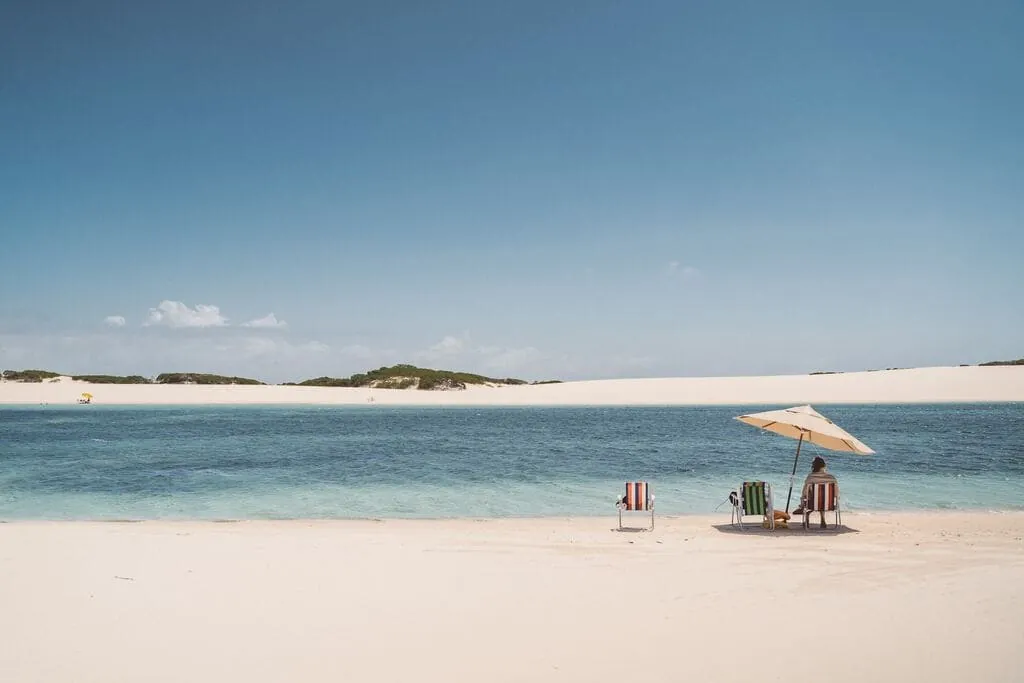 Guarda-sol e cadeiras de praia na areia branca, diante de uma lagoa de água azul com dunas ao fundo