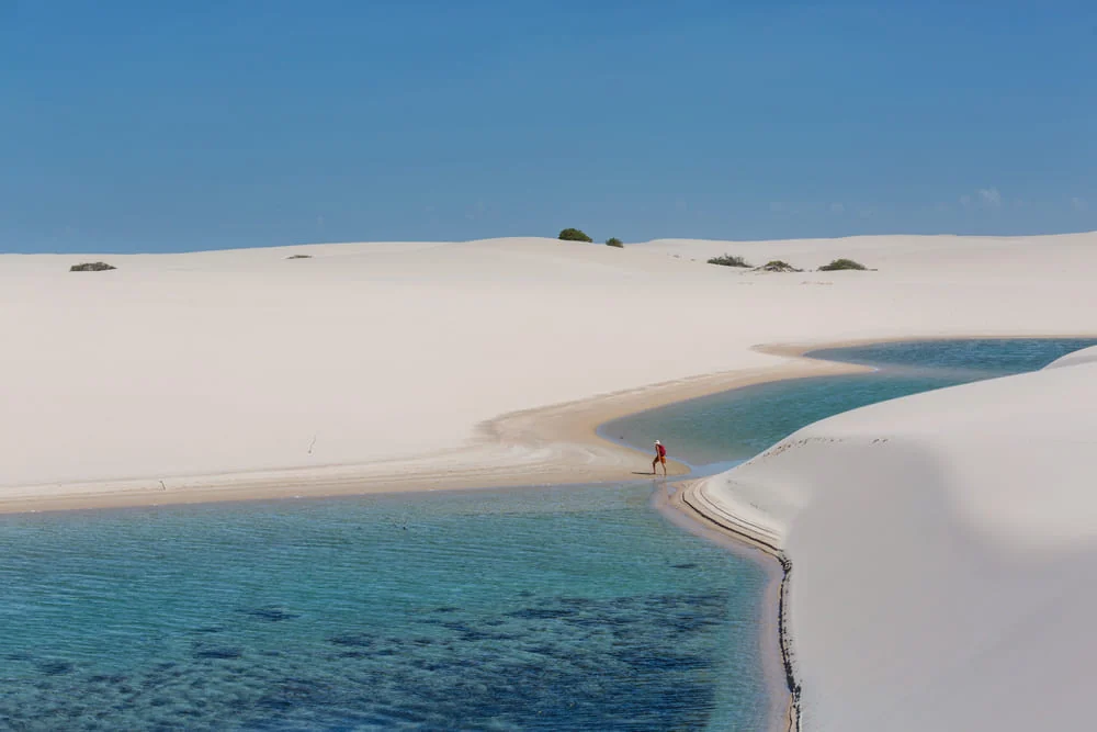 Lagoa de água azul-turquesa entre dunas brancas dos Lençóis Maranhenses com uma pessoa caminhando ao longe