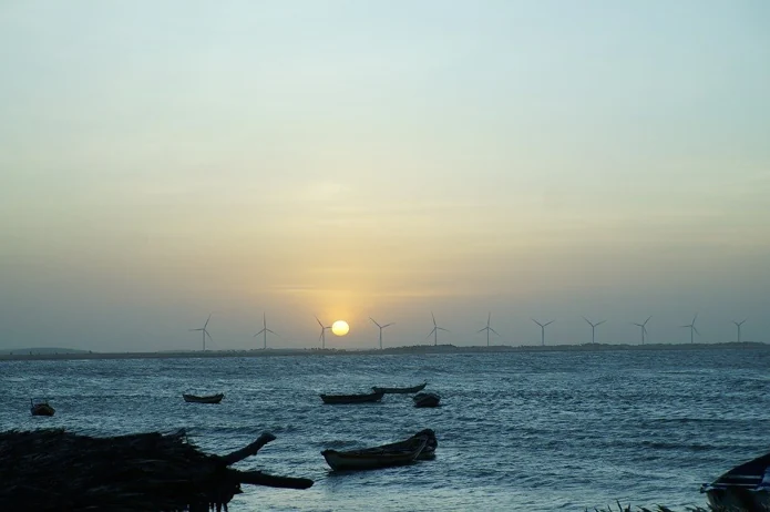Sol se pondo no horizonte atrás de uma fileira de aerogeradores, com barcos de pesca no mar em primeiro plano
