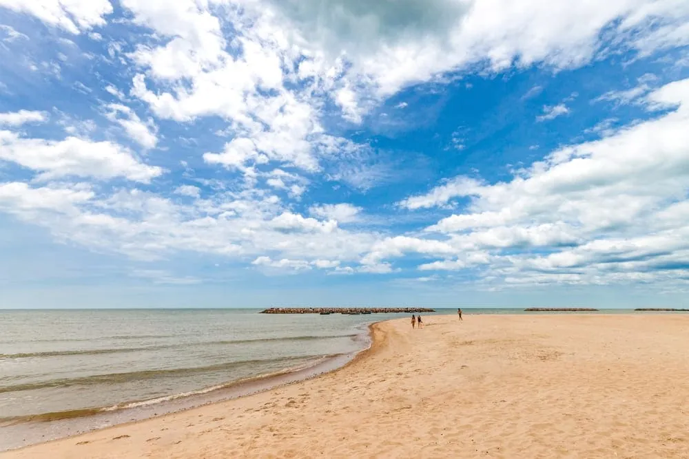 Praia de areia dourada e mar calmo, com quebra-mares de pedra ao fundo e céu azul com nuvens