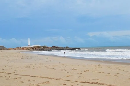 Faixa de areia e mar agitado na Praia da Pedra do Sal, com o farol branco sobre os rochedos ao fundo