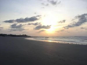 Praia ao entardecer, com o sol baixo sobre o mar e pessoas ao longe na areia