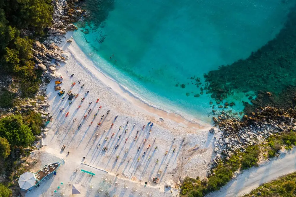 Vista aérea de praia com faixa de areia clara, mar de água turquesa, rochedos e fileiras de guarda-sóis