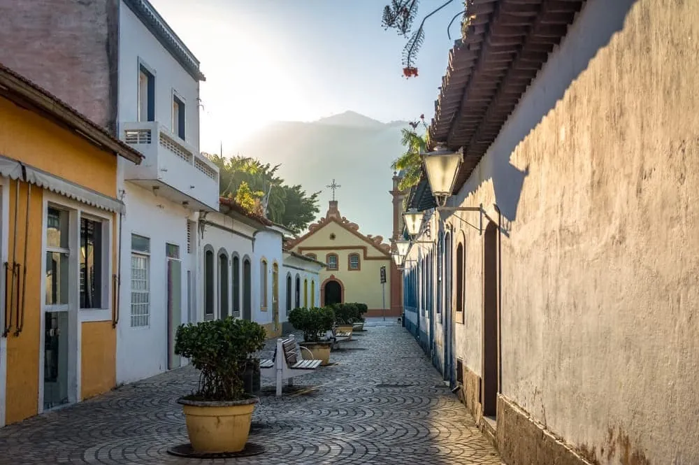 Rua de paralelepípedo ladeada por casario colonial colorido, com igreja ao fundo e lampiões nas paredes